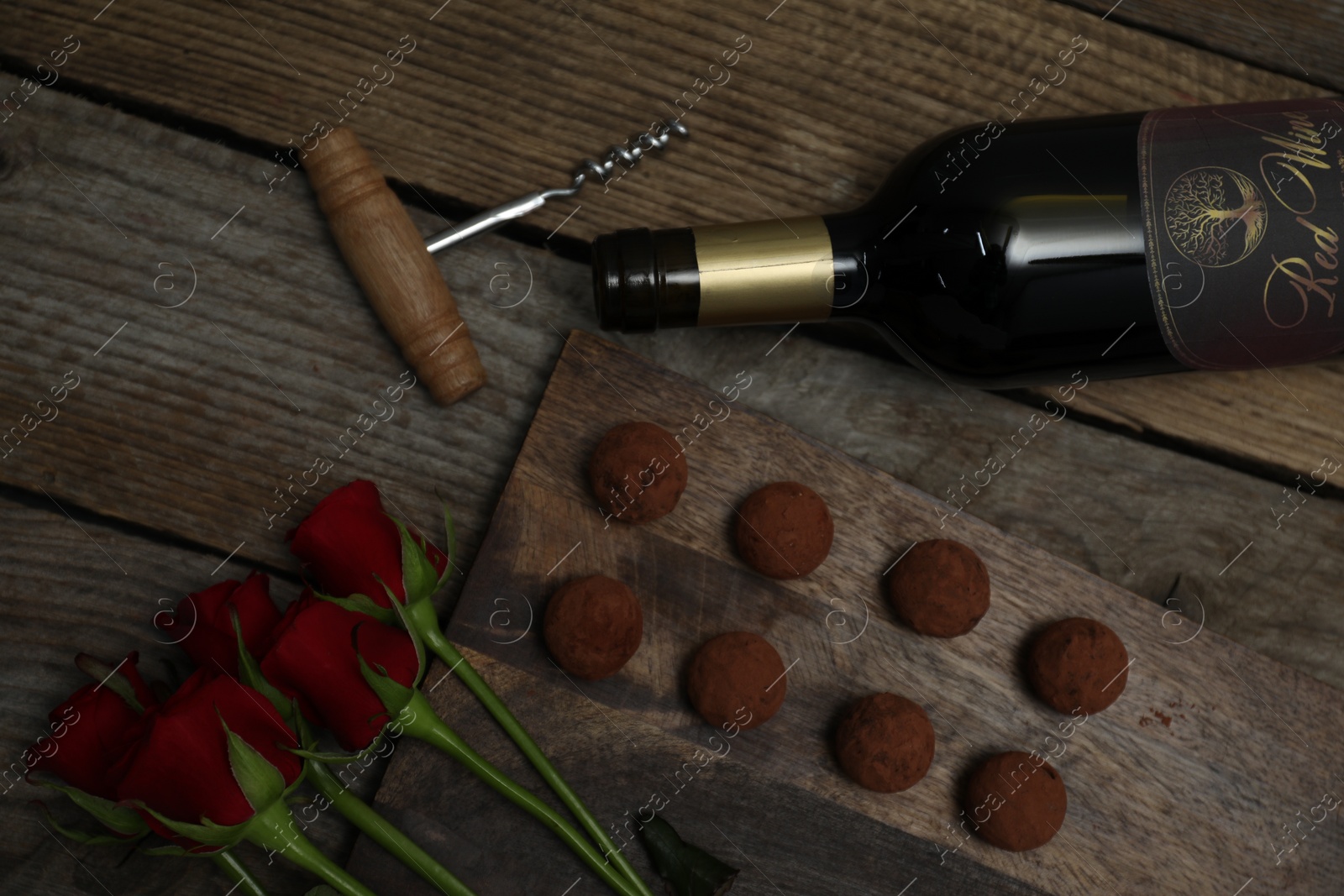 Photo of Chocolate truffles, bottle of red wine, roses and corkscrew on wooden table, flat lay