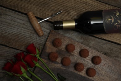 Photo of Chocolate truffles, bottle of red wine, roses and corkscrew on wooden table, flat lay