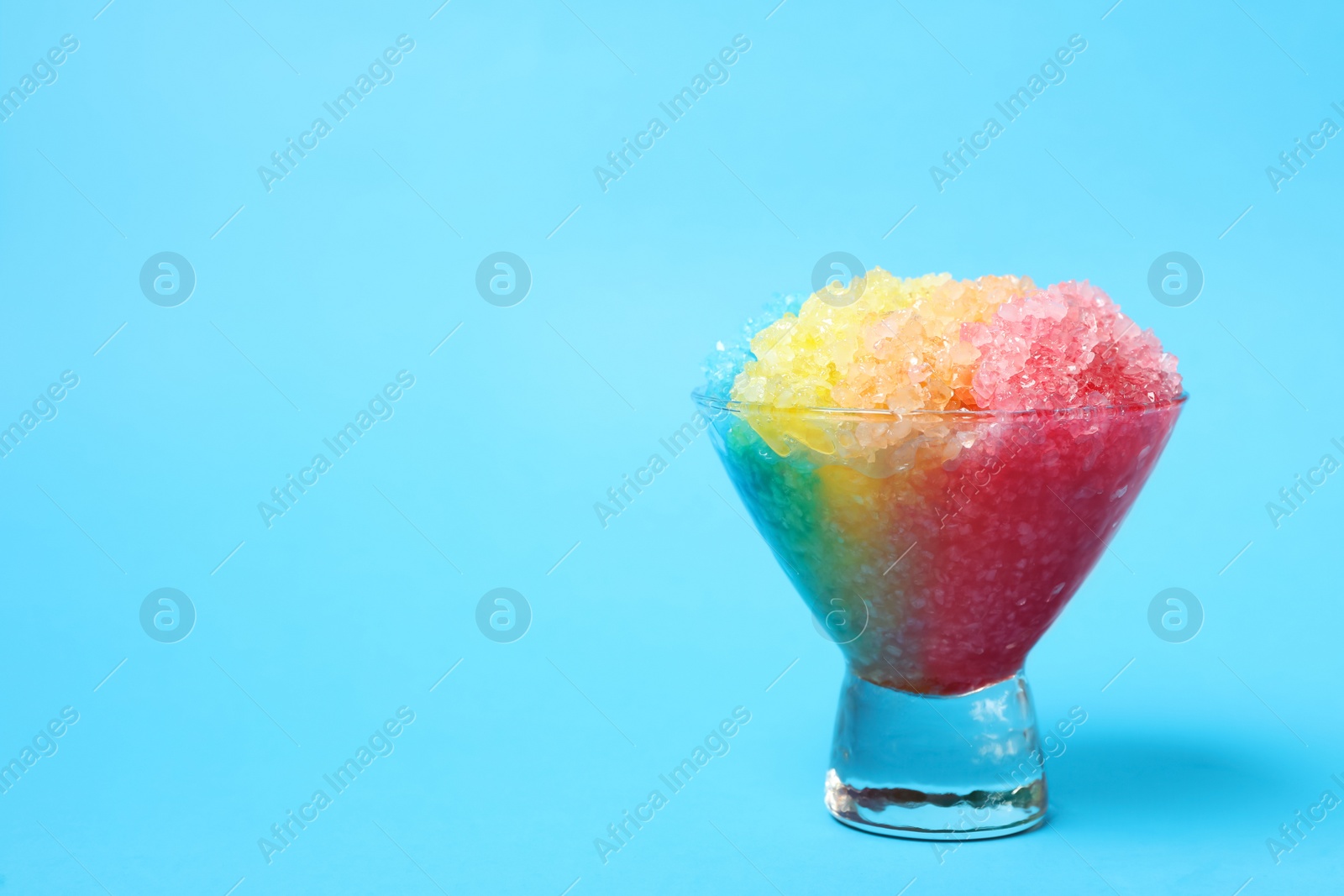 Photo of Rainbow shaving ice in glass dessert bowl on light blue background, space for text