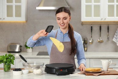 Happy woman cooking delicious crepe on electric maker at white marble table in kitchen
