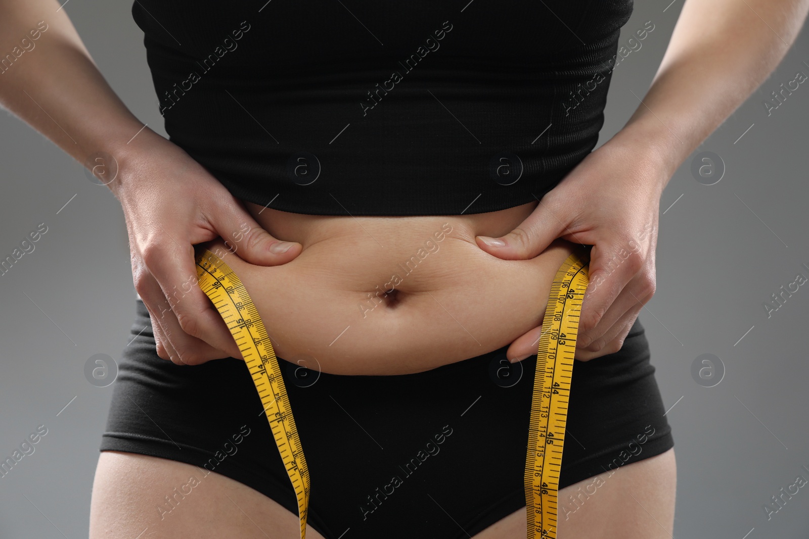 Photo of Woman with measuring tape touching belly fat on grey background, closeup. Overweight problem