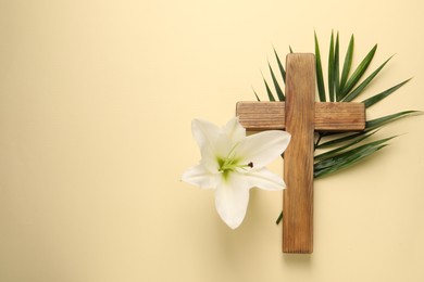 Photo of Wooden cross, lily flower and palm leaf on pale yellow background, top view with space for text. Easter attributes