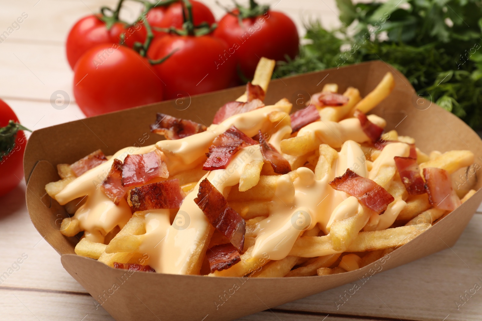 Photo of Tasty potato fries, cheese sauce, bacon and products in paper container on light wooden table, closeup