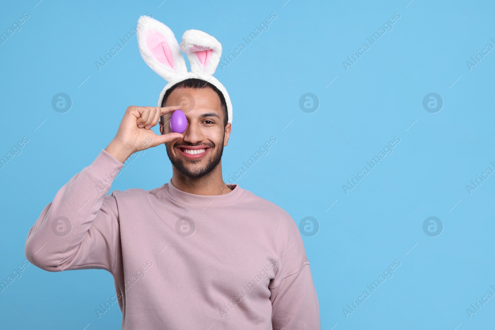 Photo of Happy African American man in bunny ears headband covering eye with Easter egg on light blue background. Space for text