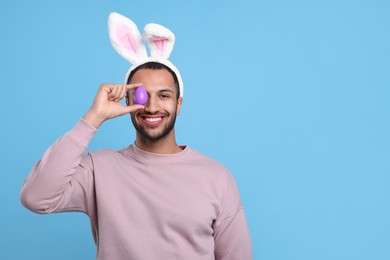 Photo of Happy African American man in bunny ears headband covering eye with Easter egg on light blue background. Space for text
