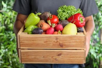 Photo of Farmer with wooden crate full of different vegetables and fruits outdoors, closeup. Harvesting time