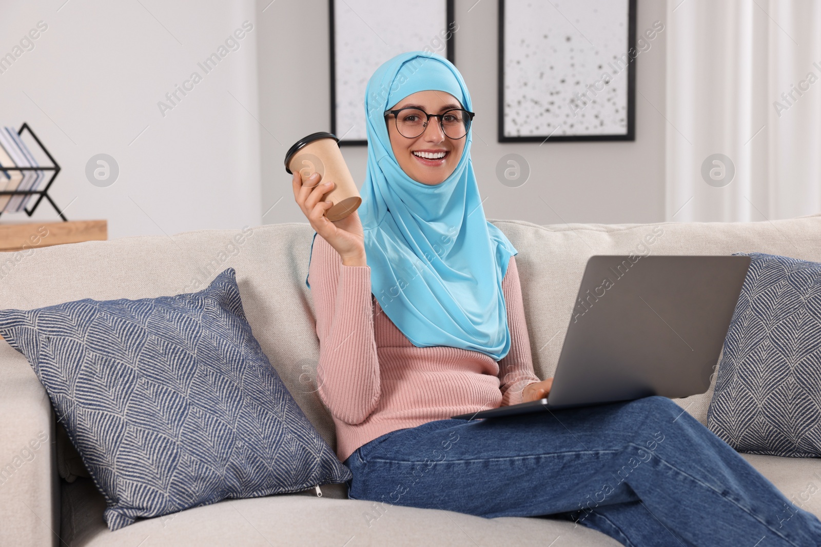 Photo of Muslim woman with cup of coffee using laptop at couch in room