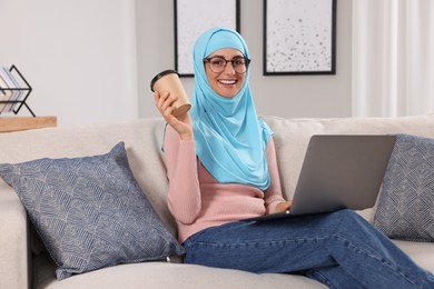Photo of Muslim woman with cup of coffee using laptop at couch in room