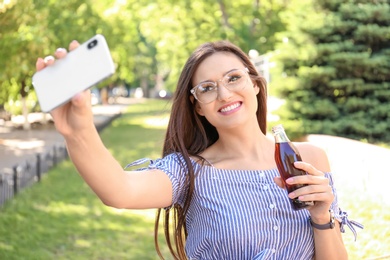 Photo of Young woman with bottle of cola taking selfie outdoors