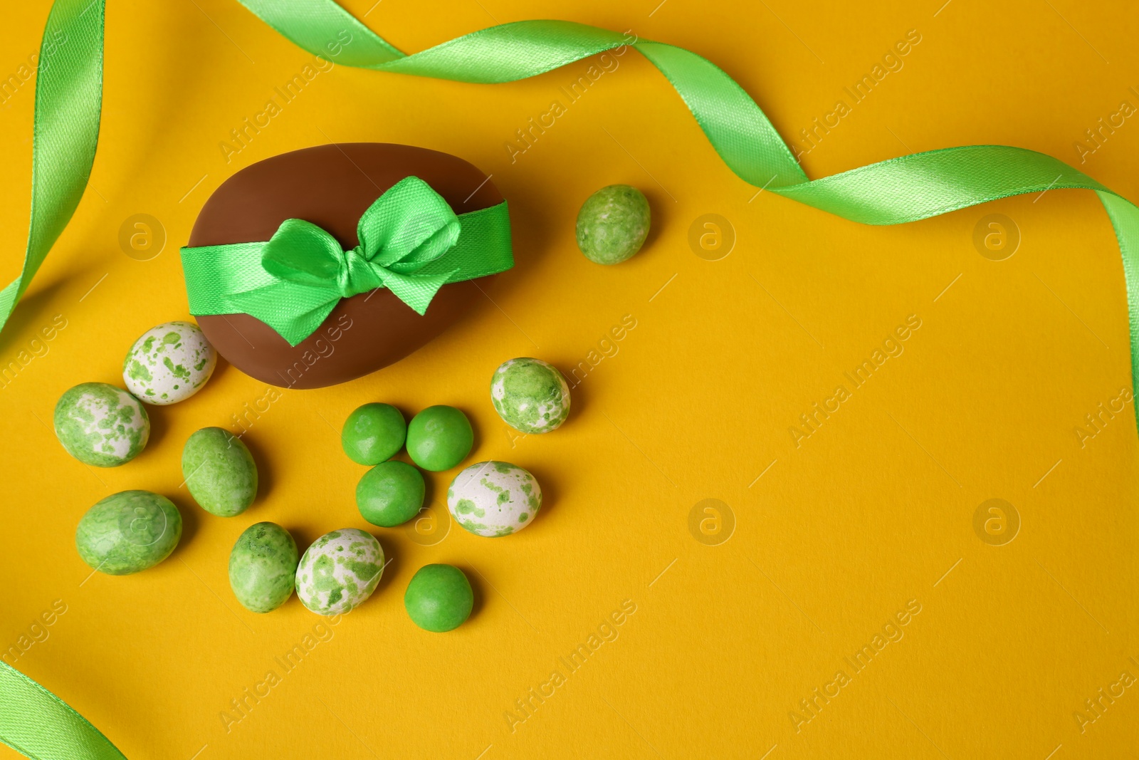 Photo of Tasty chocolate egg with green ribbon and candies on yellow background, flat lay. Space for text