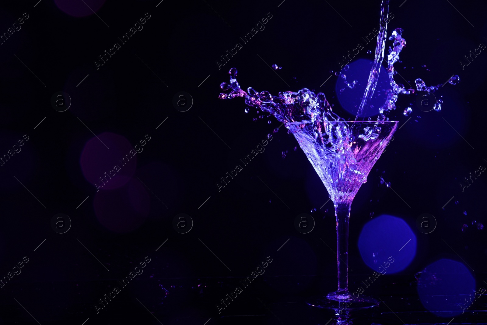 Photo of Pouring martini. Glass and splashes in neon lights on dark background, bokeh effect with space for text
