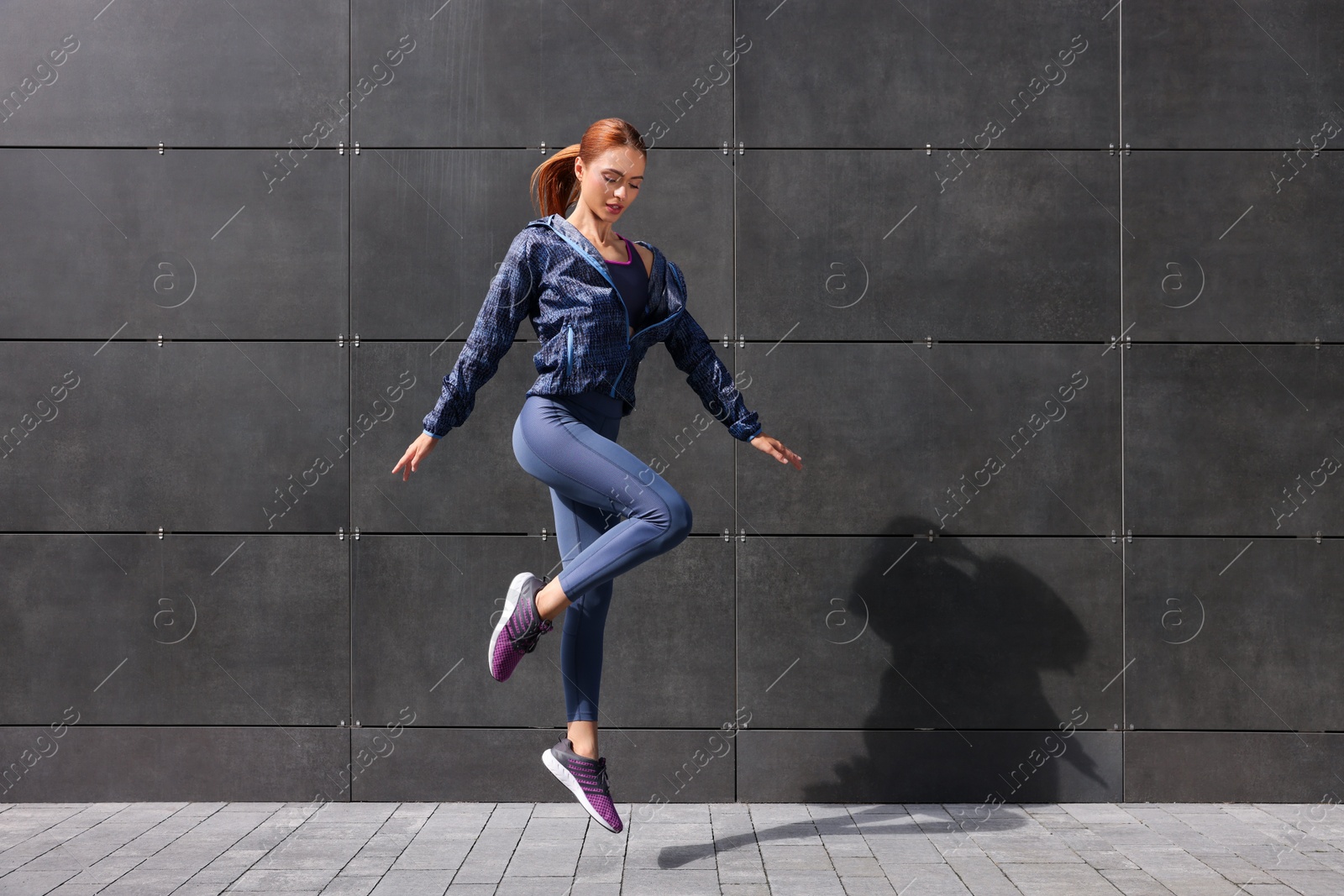 Photo of Beautiful woman in gym clothes jumping near dark grey wall on street, space for text