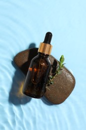 Bottle of cosmetic oil, leaves and stone in water on light blue background, top view