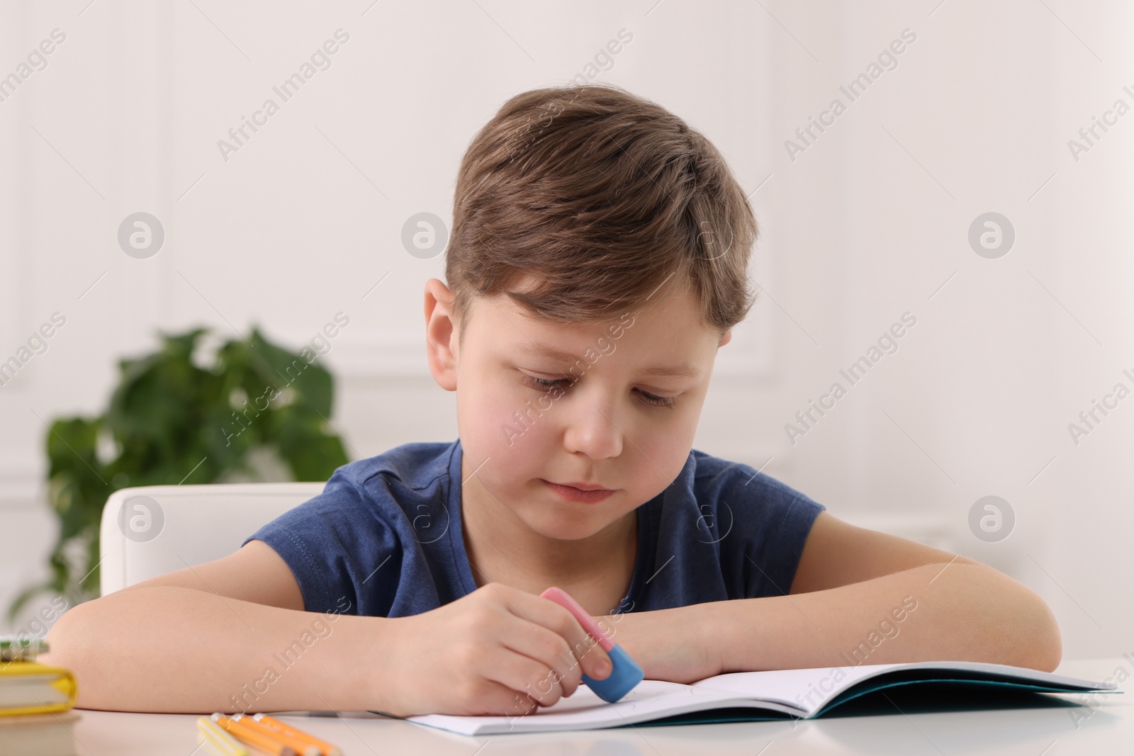 Photo of Little boy erasing mistake in his notebook at white desk indoors