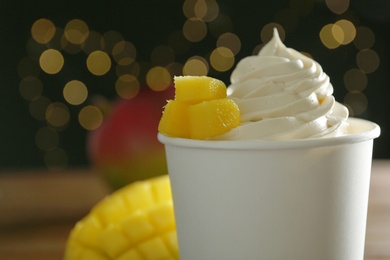 Cup of tasty frozen yogurt with mango against blurred lights, closeup. Space for text