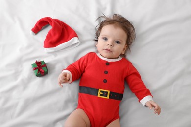 Photo of Cute baby wearing festive Christmas costume near gift box on white bedsheet, top view