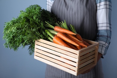 Woman holding wooden crate with ripe carrots on light blue background, closeup