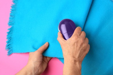 Photo of Woman cleaning light blue scarf with fabric shaver on pink background, top view