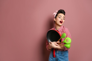 Photo of Funny young housewife with frying pan and cooking utensils on color background