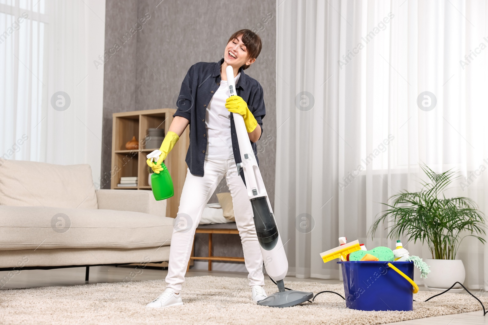 Photo of Happy young housewife with spray bottle having fun while cleaning carpet at home