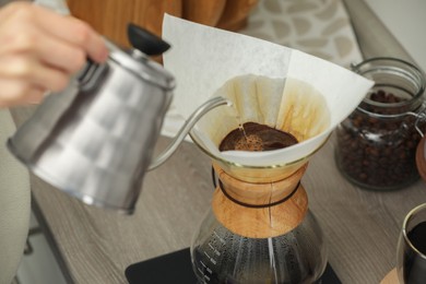 Woman pouring hot water into glass chemex coffeemaker with paper filter and coffee at countertop in kitchen, closeup