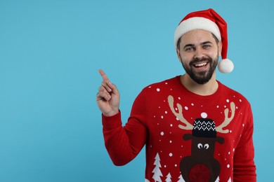 Photo of Happy young man in Christmas sweater and Santa hat pointing at something on light blue background. Space for text