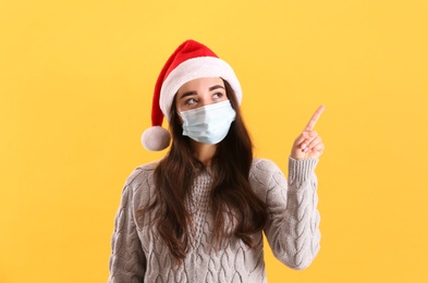 Photo of Beautiful woman wearing Santa Claus hat and medical mask on yellow background