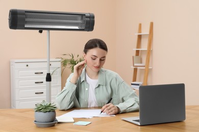 Photo of Young woman writing notes at wooden table and modern electric infrared heater indoors