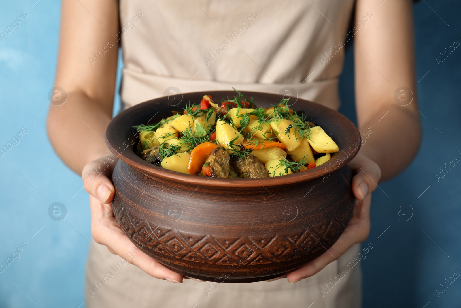 Photo of Woman holding earthenware with delicious roasted potatoes and meat on light blue background, closeup