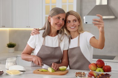 Happy daughter taking selfie with her mature mother while cooking together in kitchen