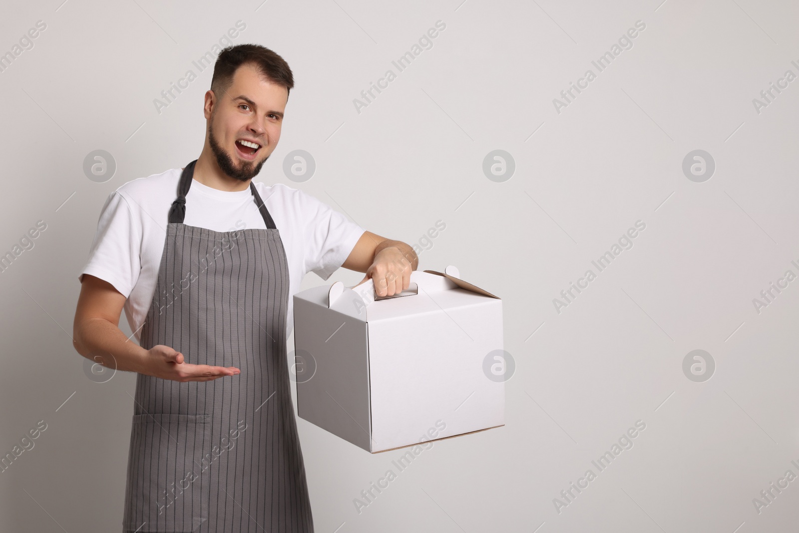 Photo of Happy professional confectioner in apron holding cake box indoors. Space for text