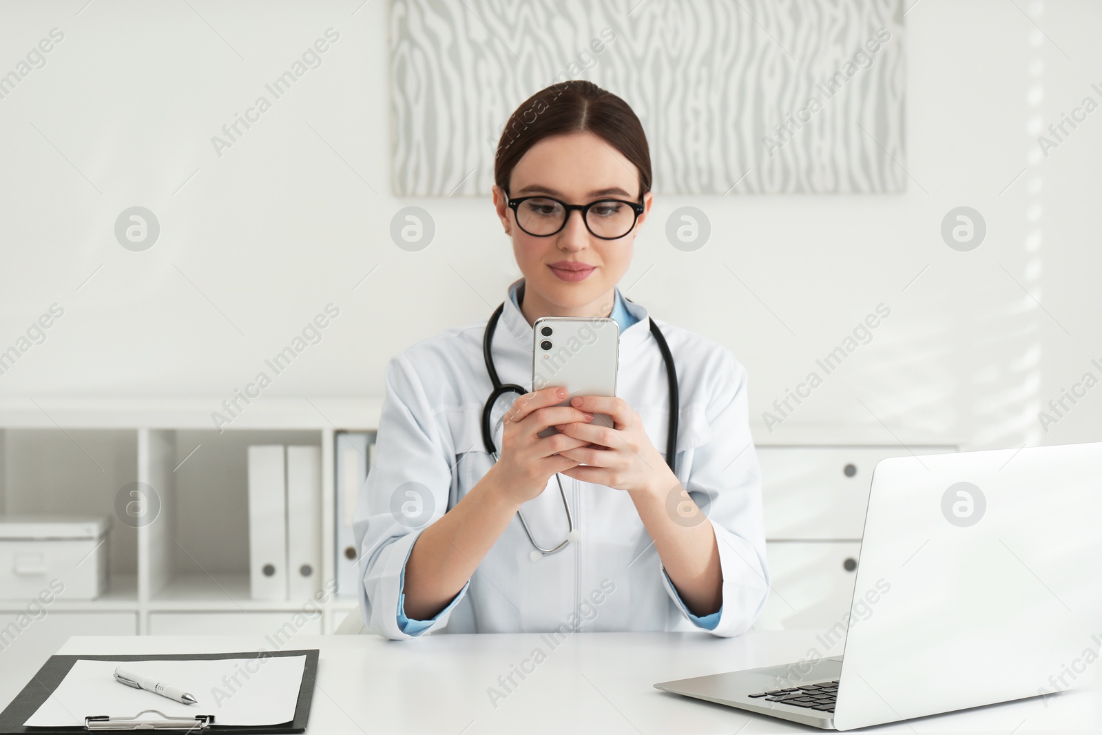 Photo of Young female doctor with smartphone at table in office