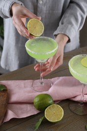Woman squeezing lime juice into glass with delicious Margarita cocktail at wooden table, closeup