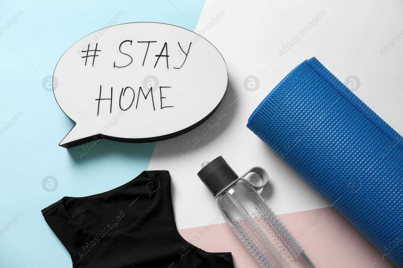 Photo of Stylish sportswear, yoga mat and speech bubble with hashtag Stay at Home on color background, flat lay. Self isolation during COVID‑19 pandemic