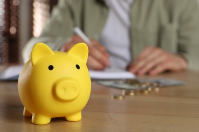 Photo of Man at wooden table, focus on yellow piggy bank. Space for text