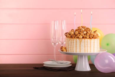 Photo of Caramel drip cake decorated with popcorn and pretzels near balloons and tableware on wooden table, space for text