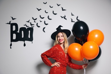 Woman in witch hat with balloons posing near white wall decorated for Halloween