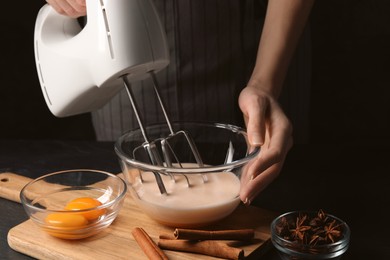 Photo of Woman whipping ingredients with mixer at black table, closeup. Cooking delicious eggnog
