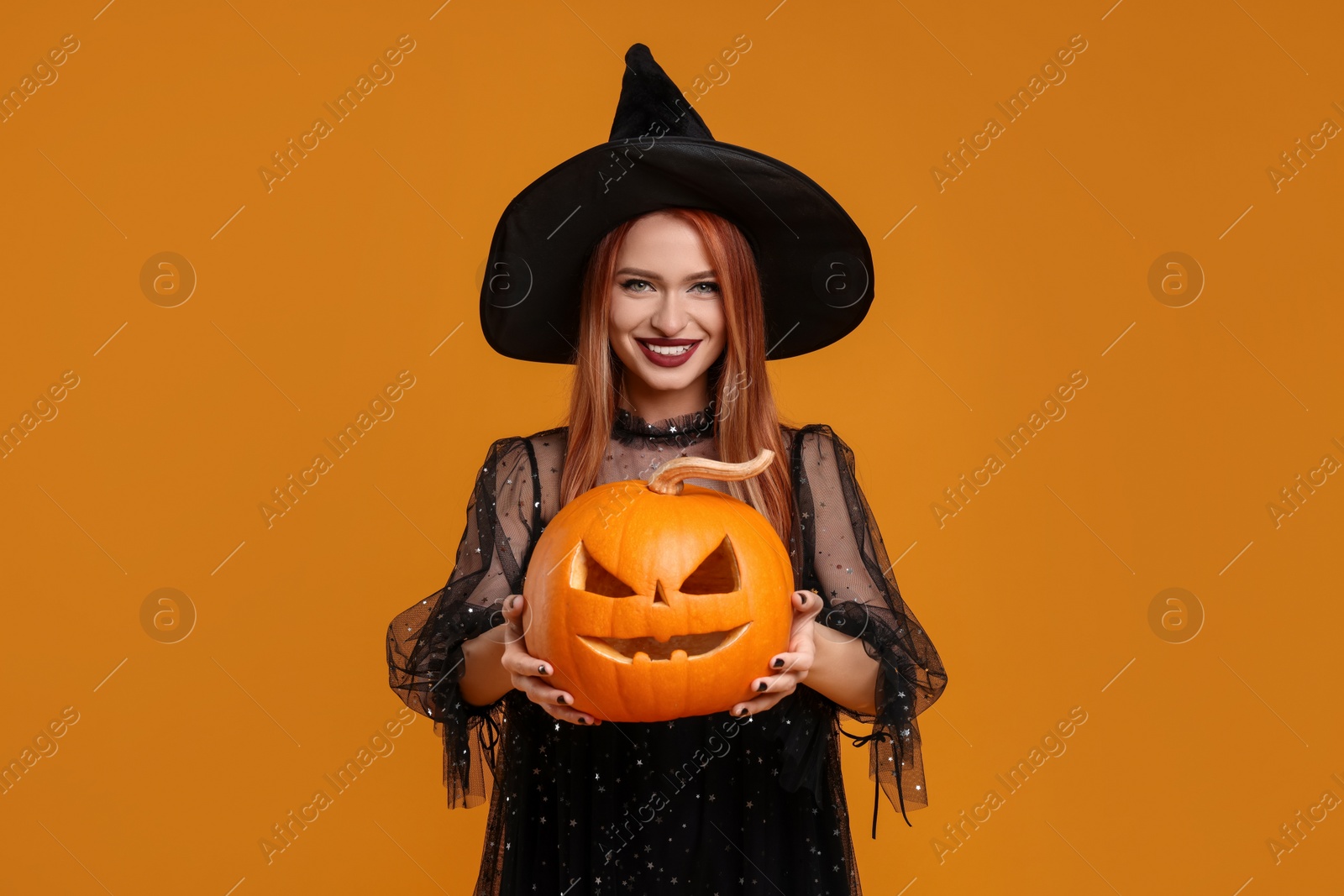 Photo of Happy young woman in scary witch costume with carved pumpkin on orange background. Halloween celebration