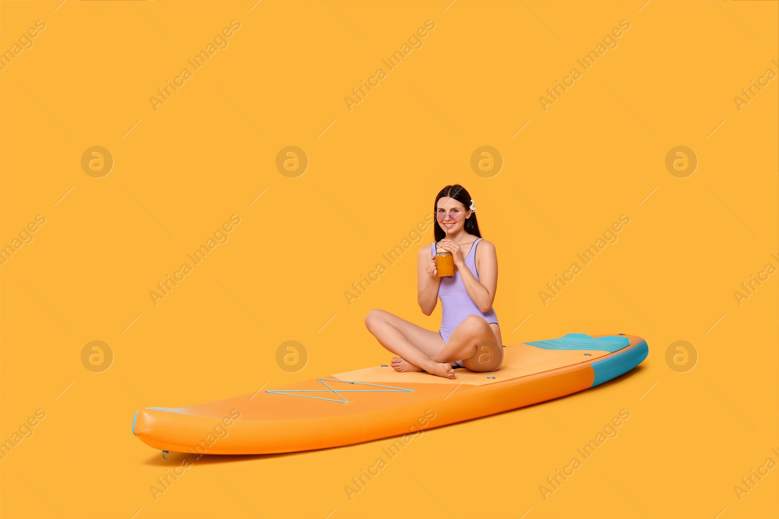 Photo of Happy woman with refreshing drink resting on SUP board against orange background