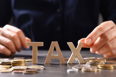 Photo of Woman with word Tax, wooden cubes and coins at grey table, closeup