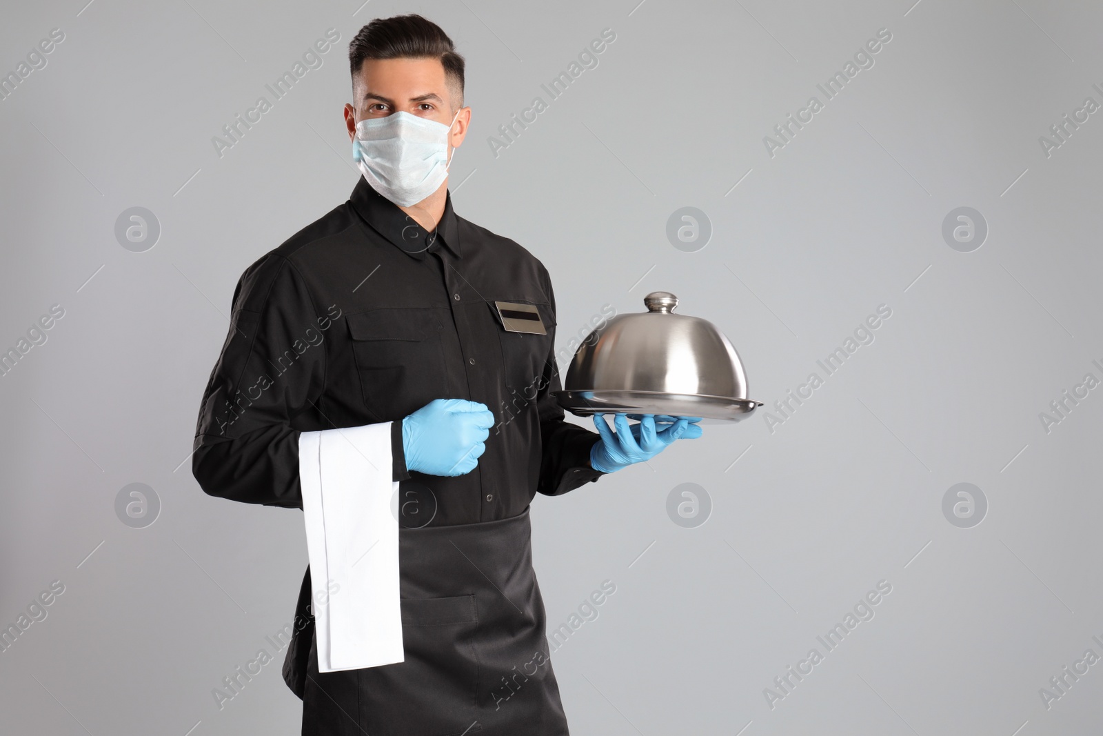 Photo of Waiter in medical face mask holding tray with lid on light grey background