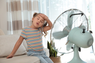 Photo of Little girl enjoying air flow from fan on sofa in living room. Summer heat