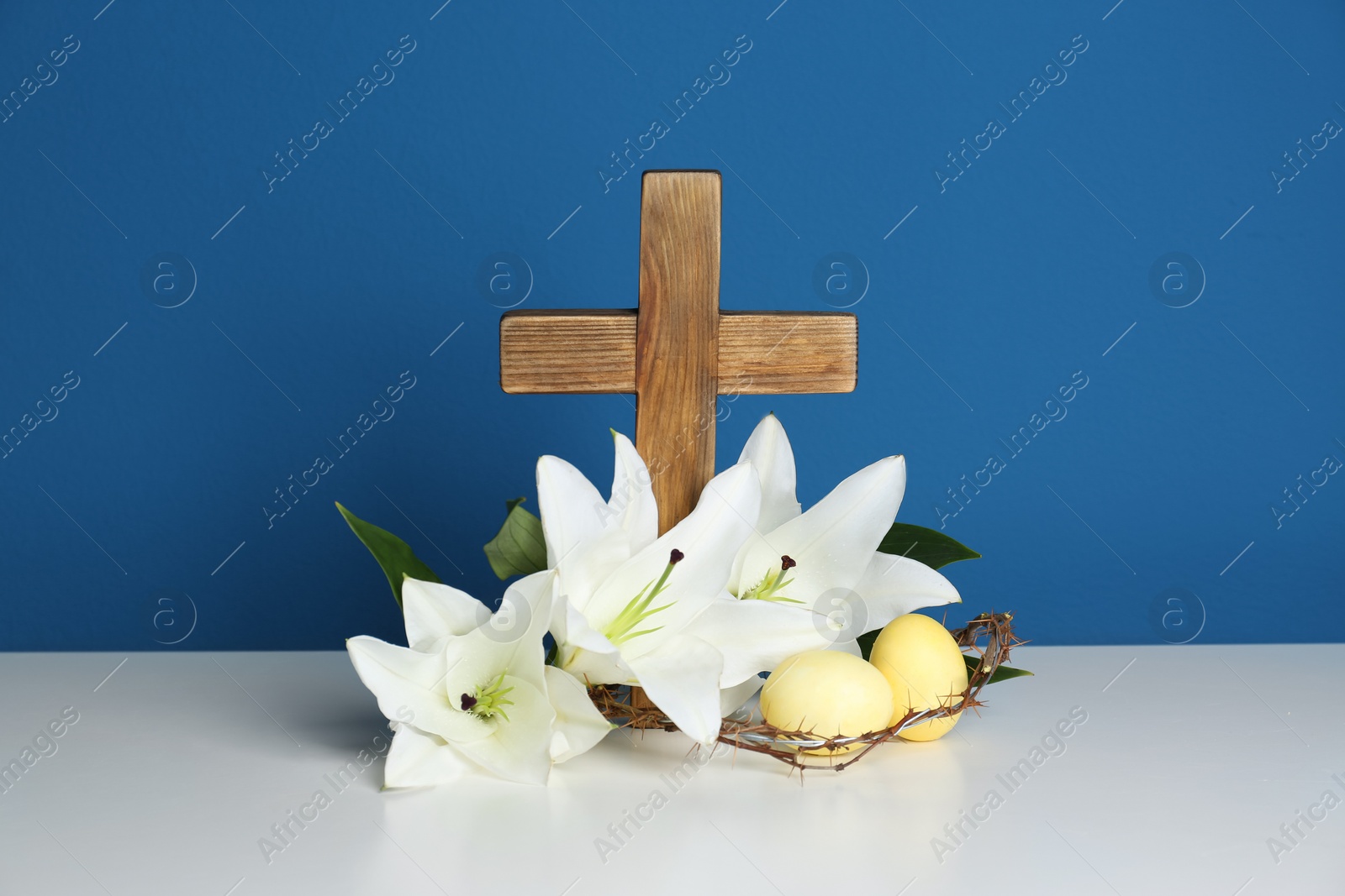 Photo of Wooden cross, crown of thorns, Easter eggs and blossom lilies on table against color background