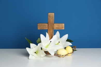 Photo of Wooden cross, crown of thorns, Easter eggs and blossom lilies on table against color background