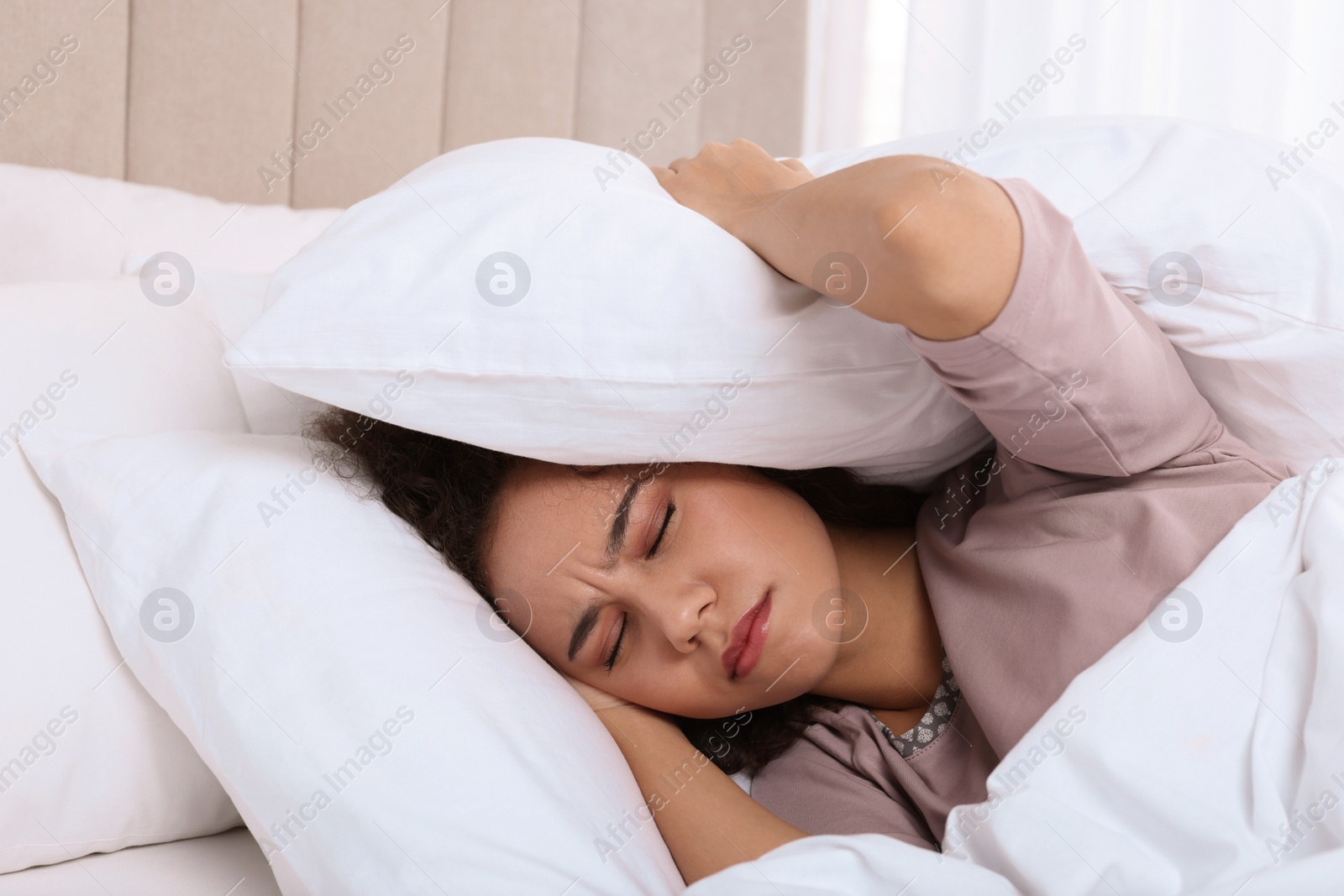 Photo of Unhappy African American woman covering ears with pillows in bed at home