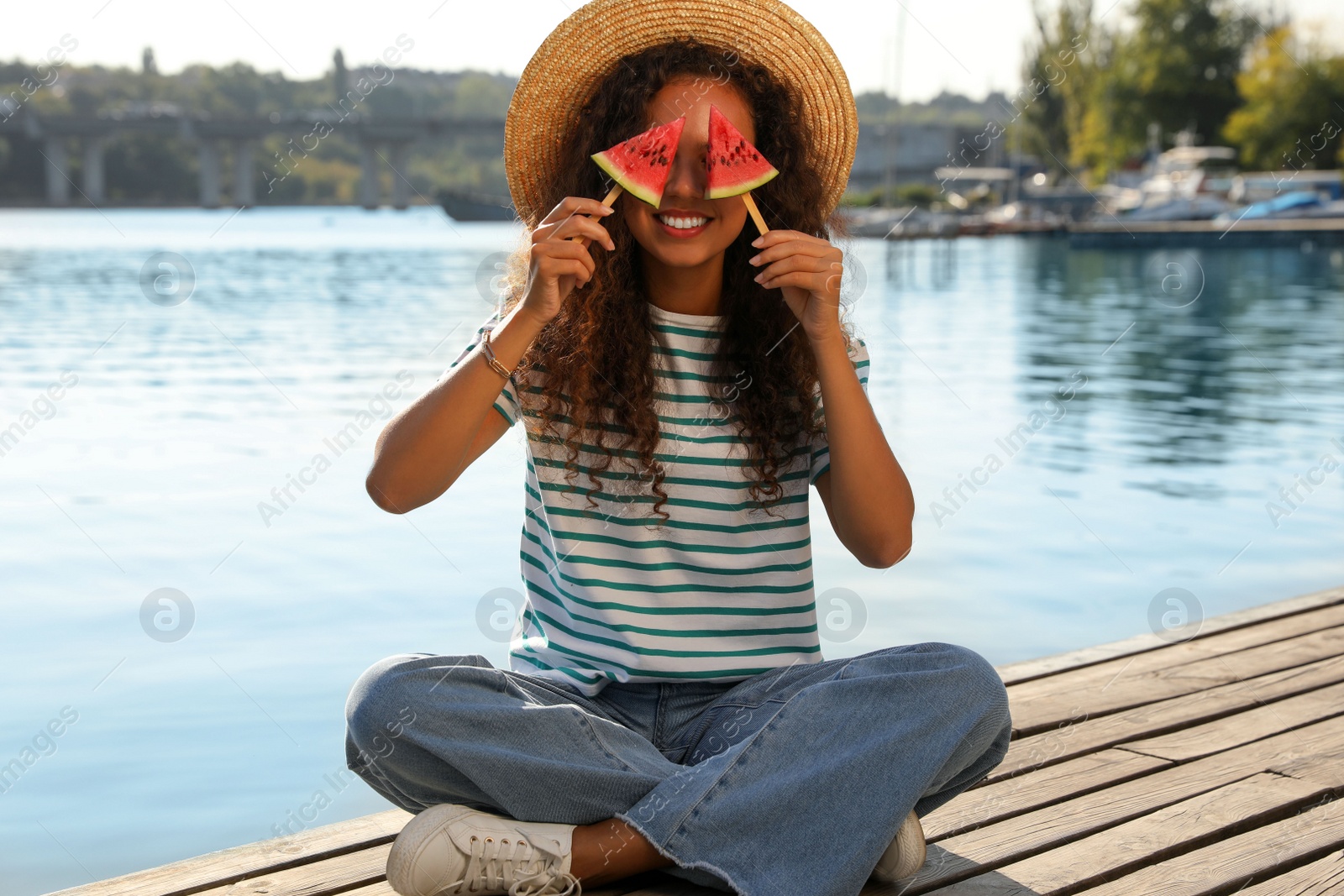 Photo of Beautiful young African American woman with pieces of watermelon on wooden pier near river