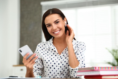 Photo of Happy young woman with smartphone listening to music through wireless earphones at table in office