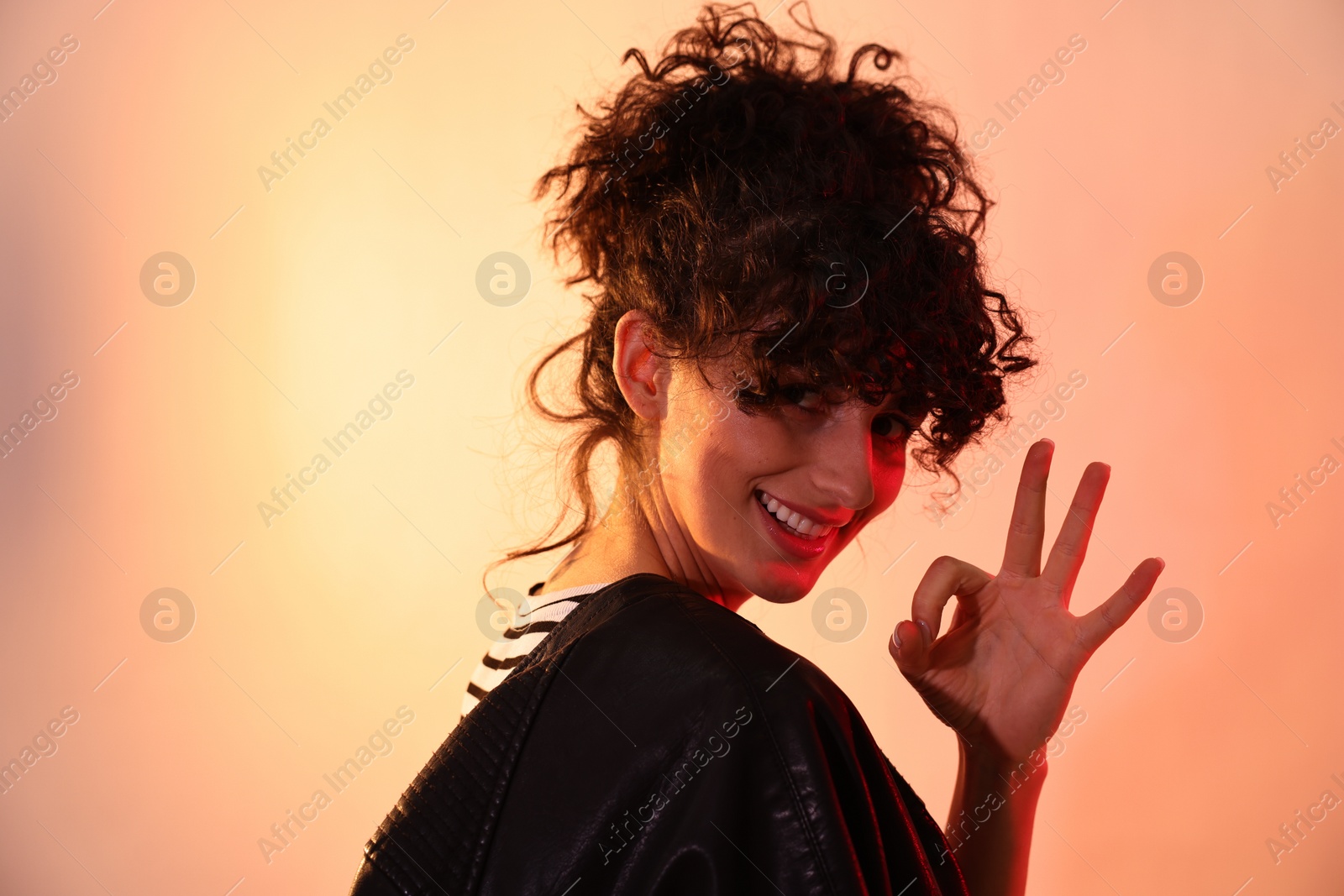 Photo of Beautiful young woman in black leather jacket showing ok gesture on color background in neon lights