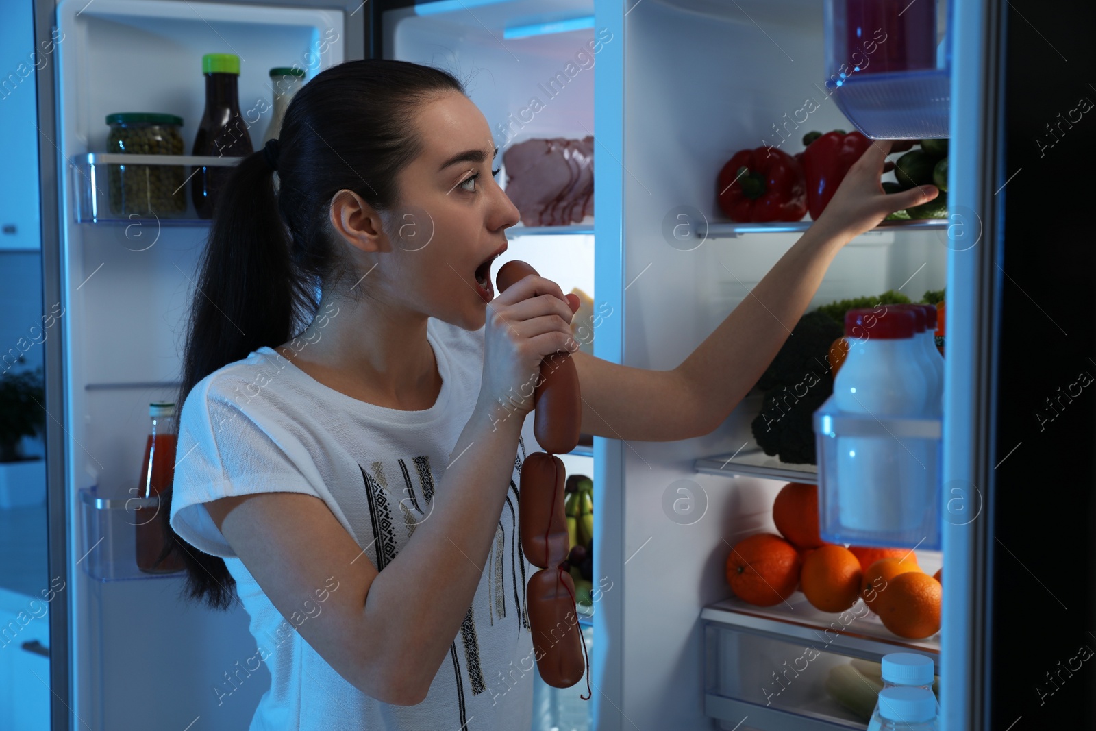 Photo of Young woman eating sausages near open refrigerator at night